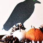 Black crow cut out with a glowing green eye set on top of a bowl of pumpkins and pinecones.