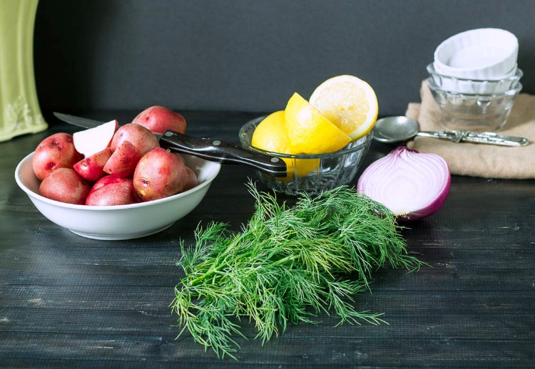 Red potatoes, wedges of lemons, fresh dill, red onion, and bowls on a table.