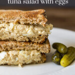 Stacked halves of a tuna egg salad sandwich with pickles on the side.