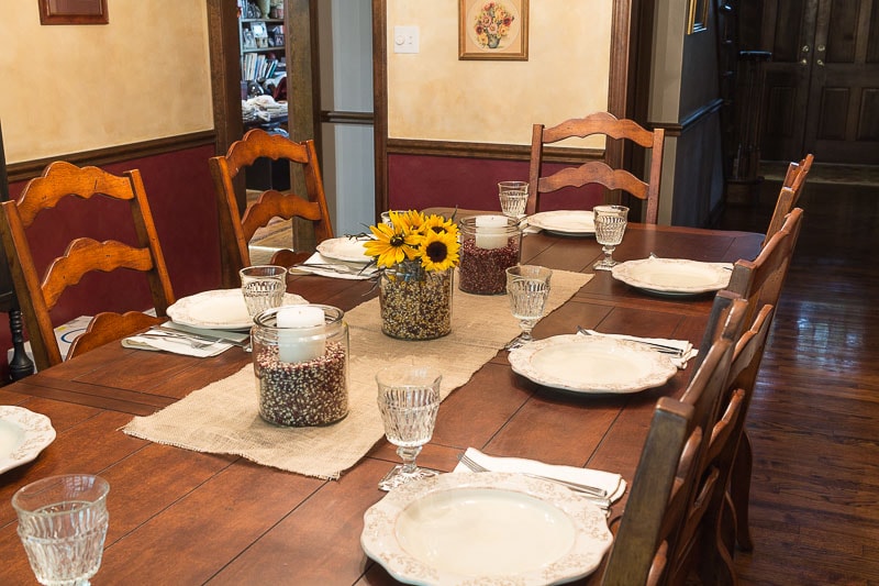 Fall table with 3 apothecary jars filled with colored popcorn, flowers and candles on a burlap runner.