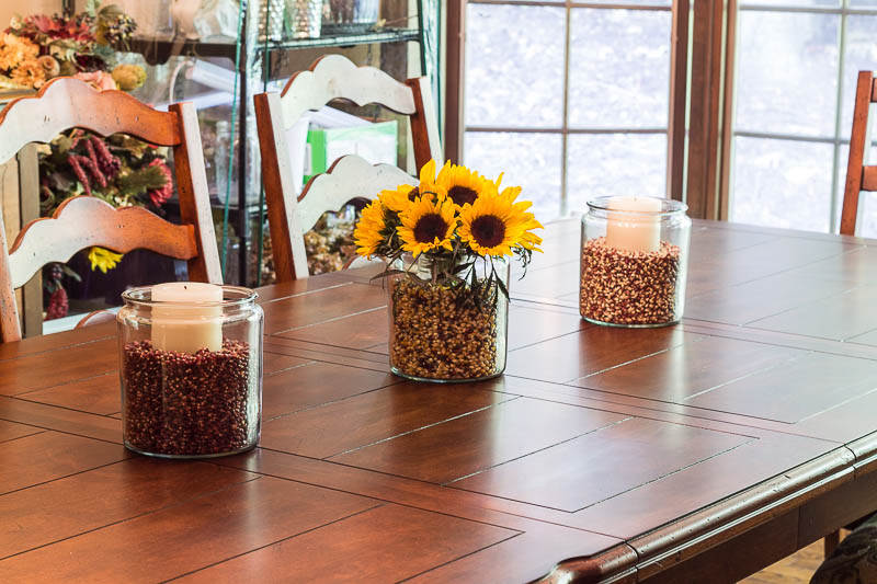 Dining room table with three apothecary jars holding color popcorn kernels, candle and one with sunflowers.