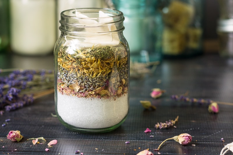 Layers of herbs and salts with a tea bag on top in a pint mason jar with scattered rosebuds and lavender around it.