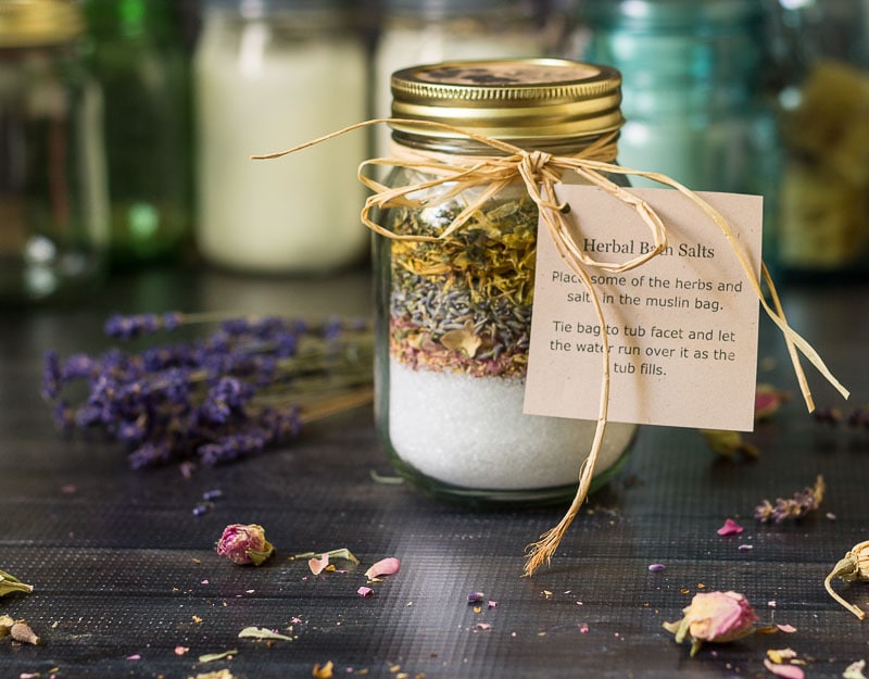 Mason jar of bath salts and layers of herbs with a raffia ribbon and instruction tag.