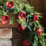 Red cardboard flowers on string of lights with greenery on a mantel.