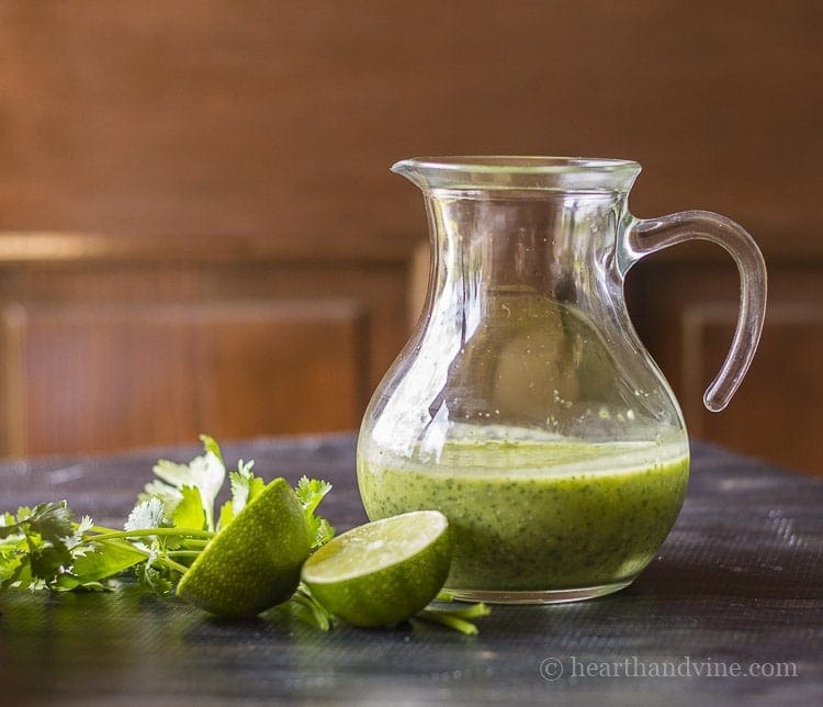 A lime cut in half, some fresh cilantro next to a small pitcher of cilantro lime dressing.