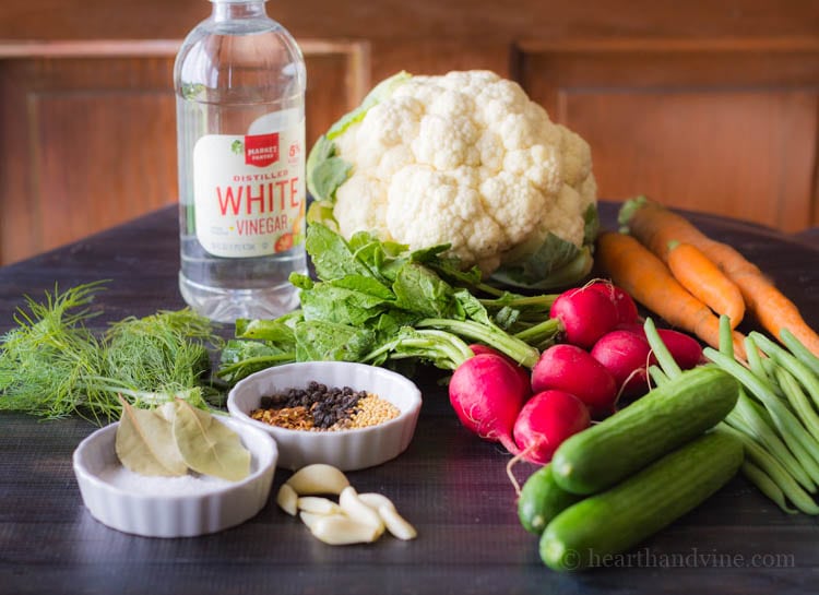 Vinegar, garlic, spices, dill, basil, bay leaves, radishes, cucumbers, green beans, carrots and cauliflower on a table.