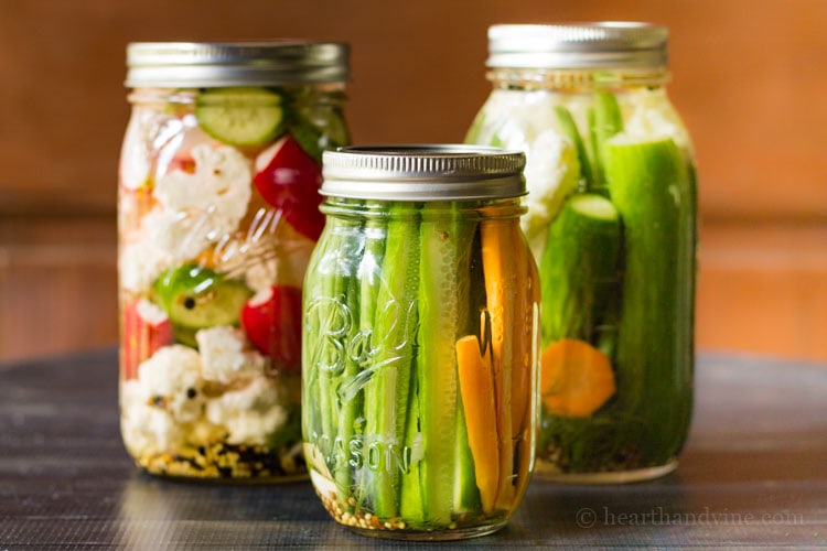 Three mason jars with pickled vegetables including carrots, green beans, cucumbers, cauliflower and radishes and whole spices.