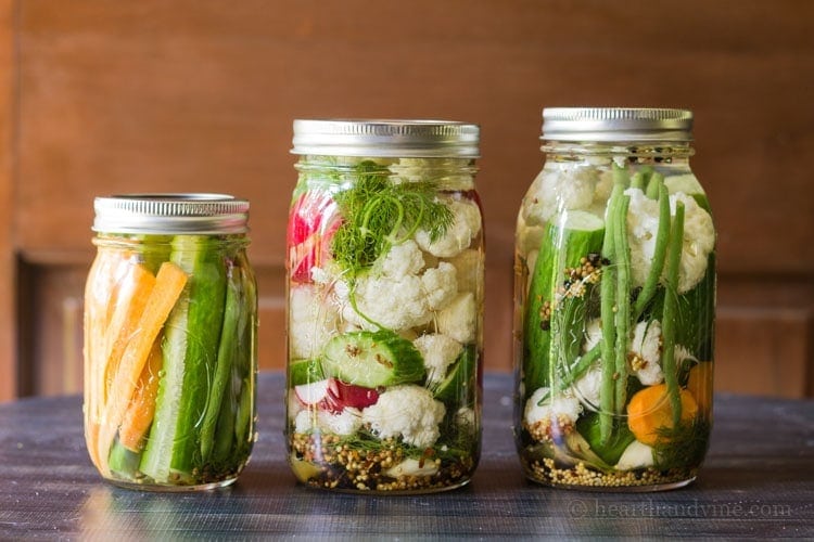 Three mason jars with pickled vegetables including carrots, green beans, cucumbers, cauliflower and radishes.