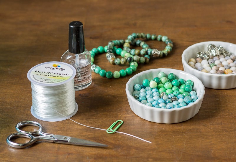 Healing stone bracelet supplies, elastic thread, stones, scissors, a paperclip and clear nail polish.