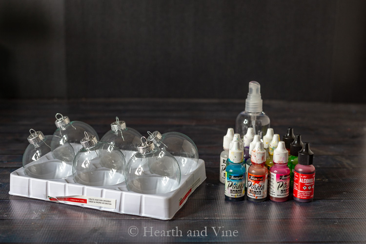 Alcohol ink ornament supplies including a dozen of plastic ball ornaments, several bottles of alcohol inks and a spray bottle with isopropyl alcohol.