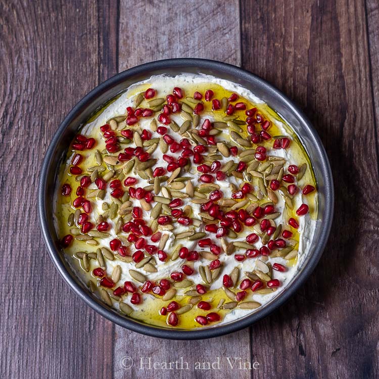 Pan of goat cheese dip with lemon olive oil, pomegranate seeds and pepita seeds on top.