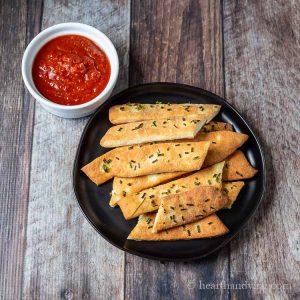Aerial view of chive breadsticks on a plate next to a bowl of marinara sauce.