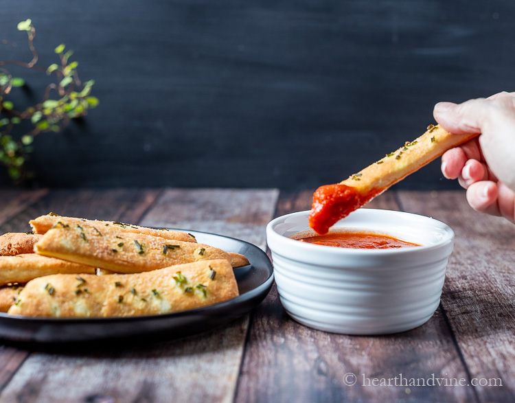 Breadstick dipped in marinara sauce on right and plate of breadsticks on left.