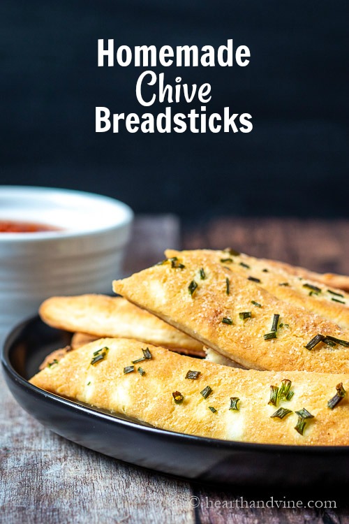 Plate of golden chive breadsticks with a partial view of a small bowl of marinara sauce above and text overlay saying homemade chive breadsticks.