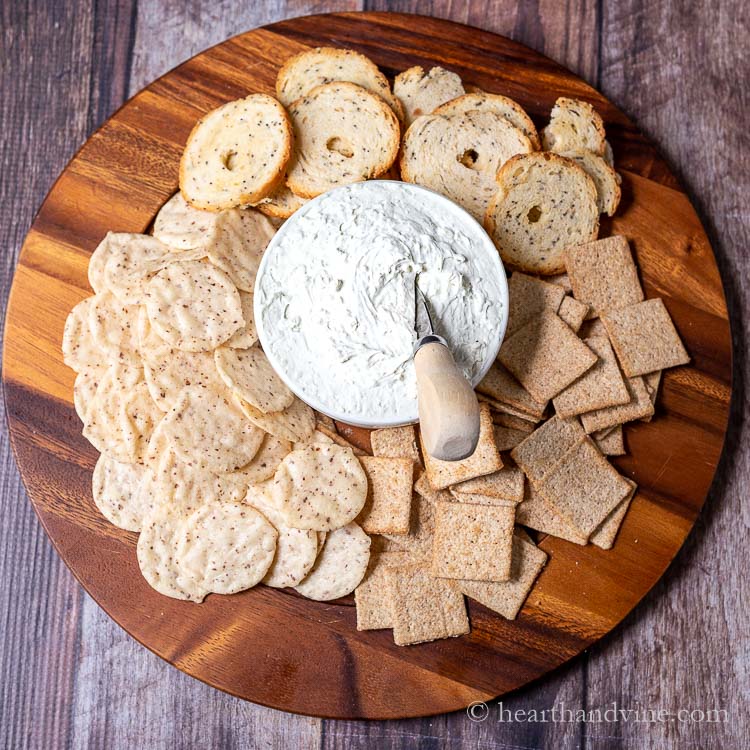 Wooden tray with crackers and bagel chips surrounding a white bowl of blue cheese spread with a small knife