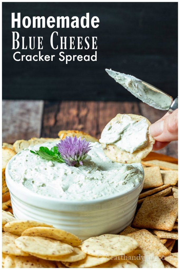 Crock of blue cheese spread surrounding by crackers on a tray. Hand with knife holding cracker with spread on it.