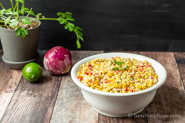 Bowl of Mexican street corn with red onion, a lime and pot of parsley next to it.