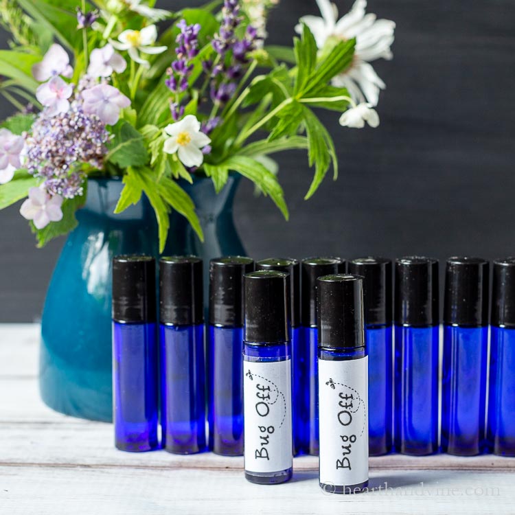 Vase with flowers behind a line of homemade insect repellent oil in cobalt blue bottles behind two bottles with a bug off label.