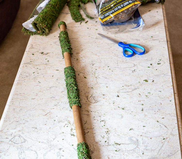 Sheet moss attached on the wooden broom stick in areas, and scissors on table.