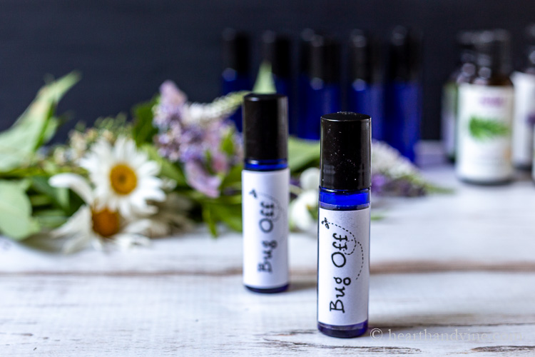 Two blue bottles with bug off labels, flowers and more plain blue bottles and essential oils in background.