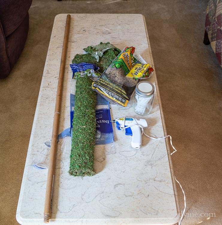 Old broom stick, sheet moss, glue gun, glue sticks on a table.