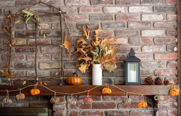 Right side of a rustic fall mantel showing pine cones, a black lantern, a white vase with oak leaves a faux green pumpkin and a branch window. Hanging from the mantel is a multi-colored twine pumpkin garland.