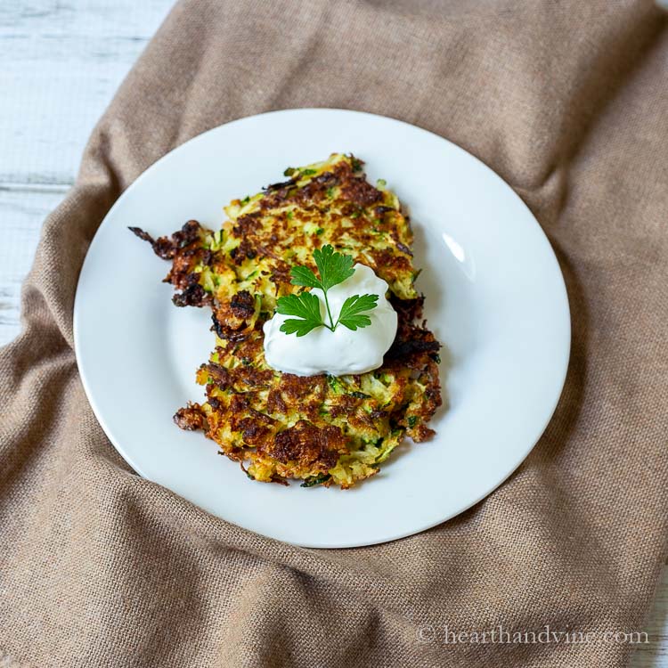 Two zucchini latkas on a plate topped with sour cream and parsley