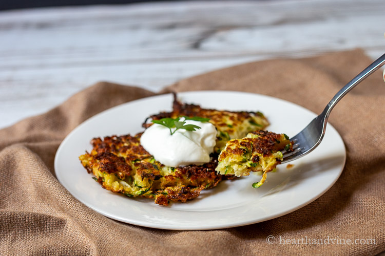 A forkful of zucchini pancake from a plate with two topped with sour cream.