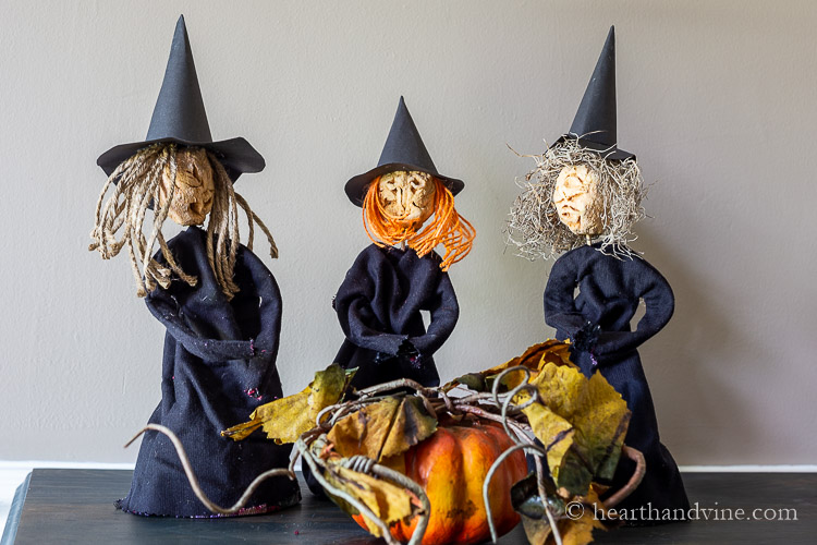 Three dried apple dolls dressed as witches on a table around a pumpkin.