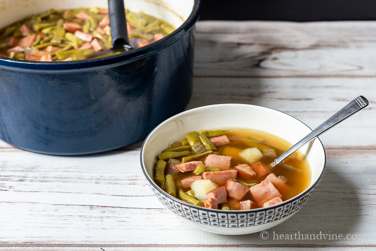Ham and string bean soup in a serving bowl next to an entire pot of soup.