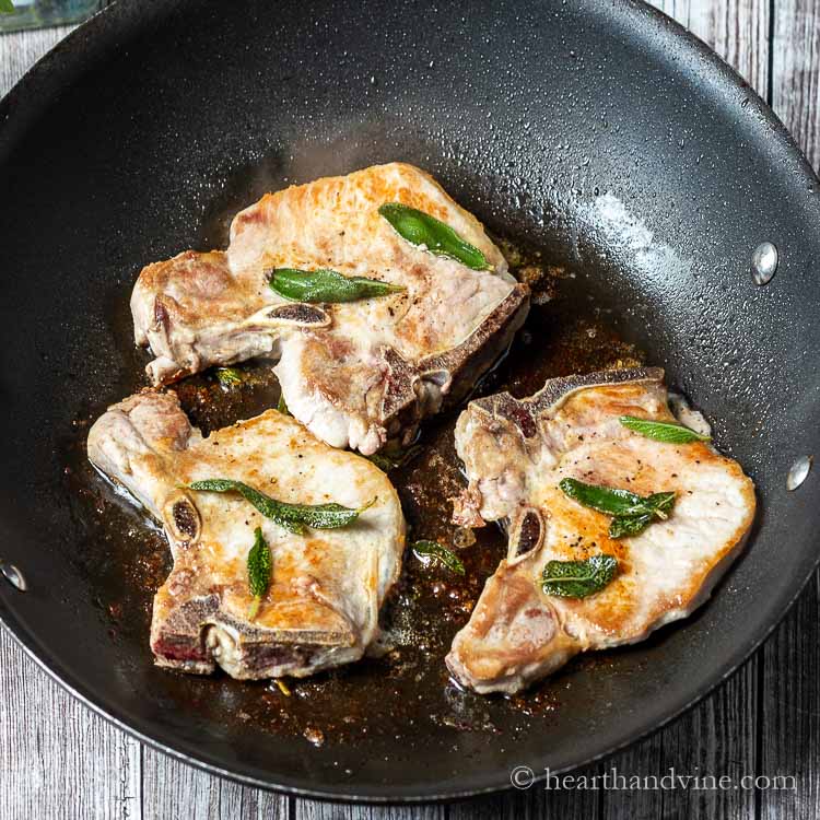 Three pork chops in a skillet with sage leaves.