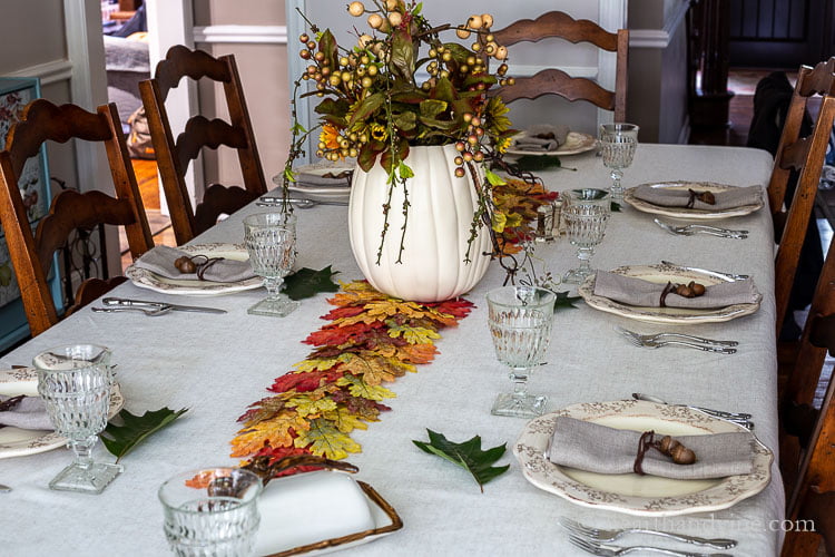 Decorated Thanksgiving tablescape showing neutral tablecloth, plates and napkins and a color center with a autumn leaf runner, and a colorful centerpiece of fall colored flowers and vines in a white pumpkin.