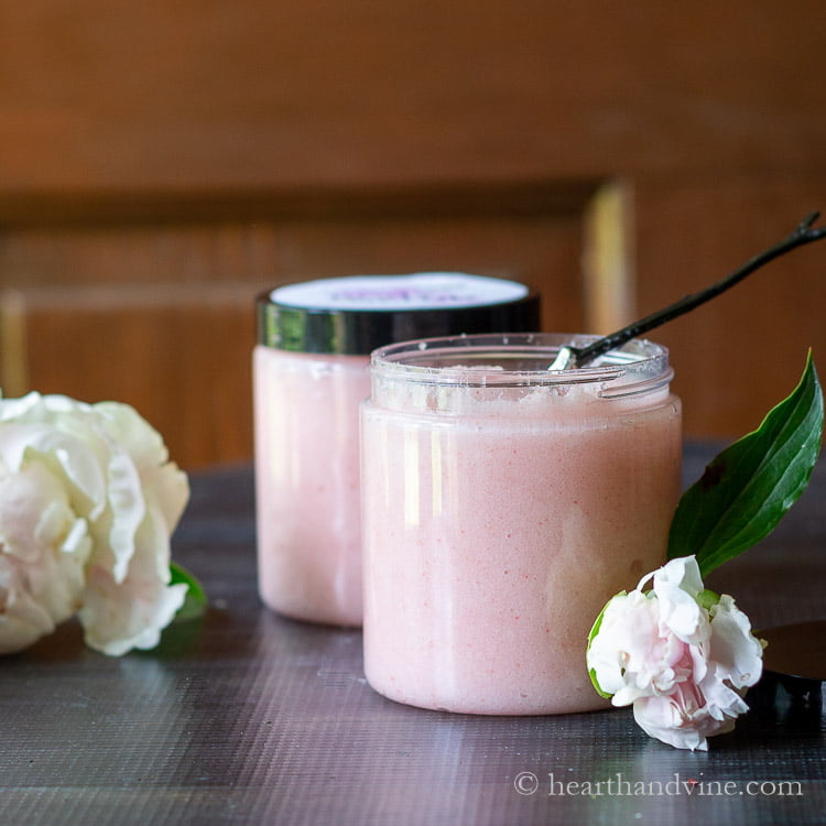 Two jars of pink body scrub with fresh peonies and a branch spoon.