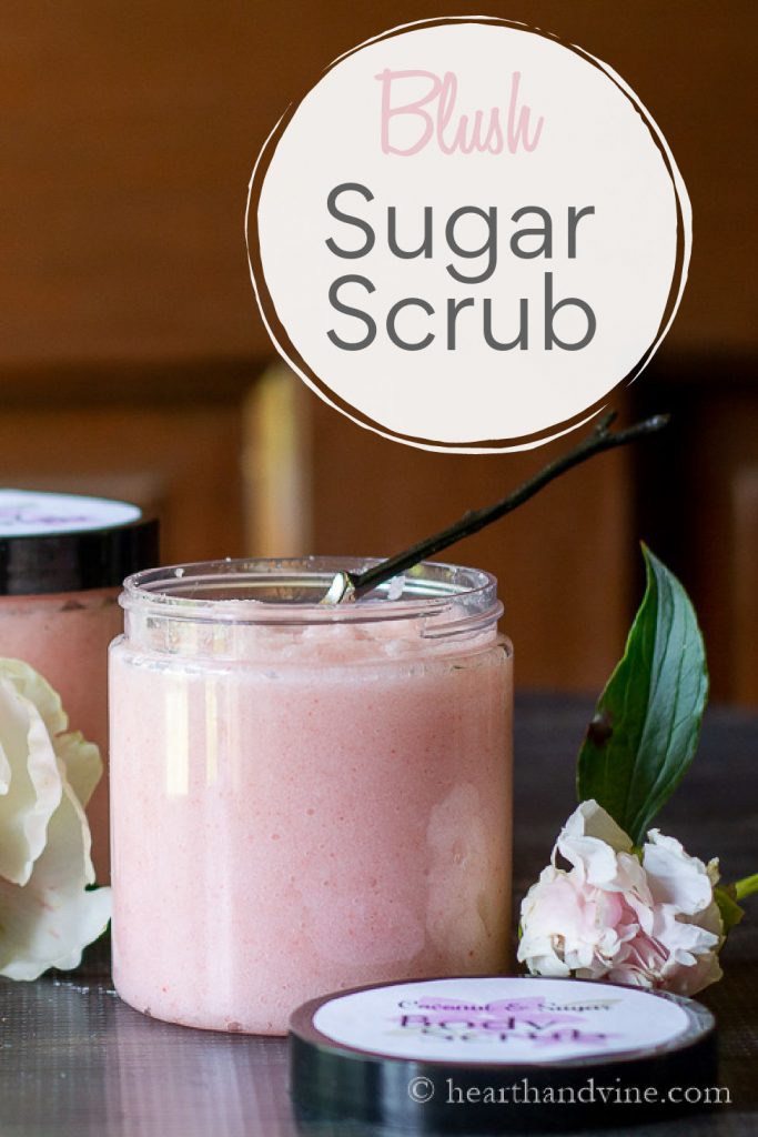Open jar of pink body scrub with peony flower bud next to it and a small spoon inside.