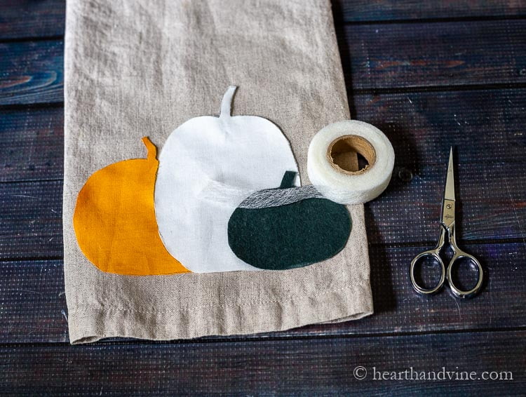 Cut out appliques of pumpkins on a linen tea towel. A roll of stitch witchery on top and a pair of small scissor on the side.