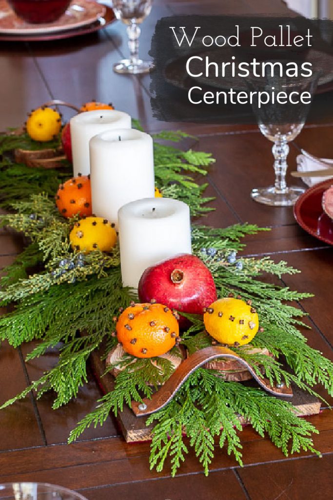 Table with a wood pallet centerpiece with white candles, cedar branches, orange & lemons studded with cloves and pomegranates.
