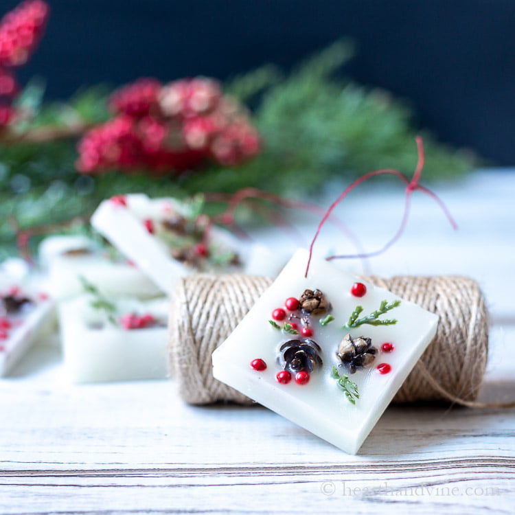 Square wax ornament with min pinecones, red berries and fake greenery leaning on a roll of twine.