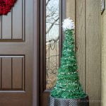 Faux Christmas tree in an outdoor planter with a white pom pom on top.
