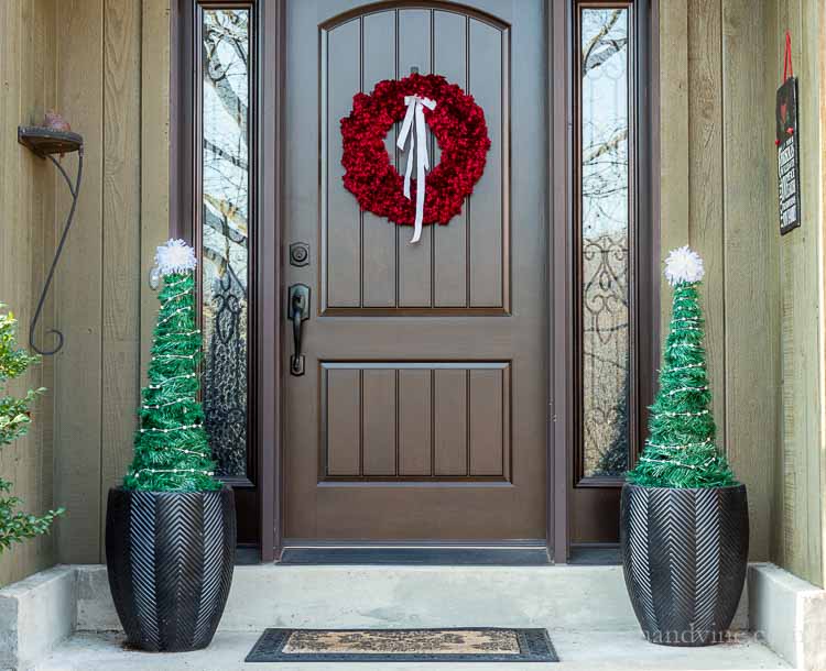 Front porch decorated for Christmas. A bright red yard wreath is on the door. Flanking the door are two black planters on each side with decorated faux Christmas trees.