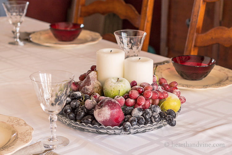 Sugared grapes, apples and pears on a glass serving tray with two white pillar candles in the middle on a dressed table.