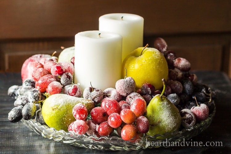 Glass tray with two white candles surrounded by sugared fruit.