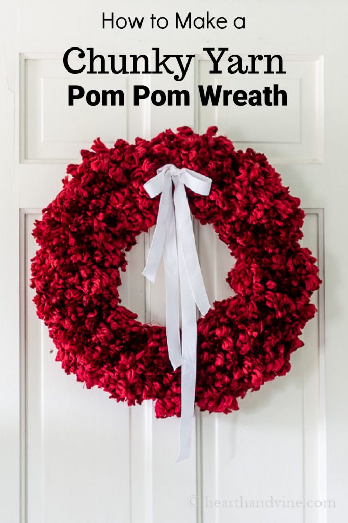 Red chunky yarn pom pom wreath on a white door with a white ribbon.
