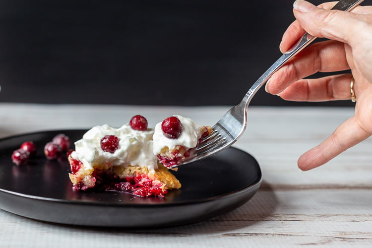 A hand taking a forkful of cranberry dump cake from a black plate with a piece of cake with whipped cream and sugared cranberries on top.