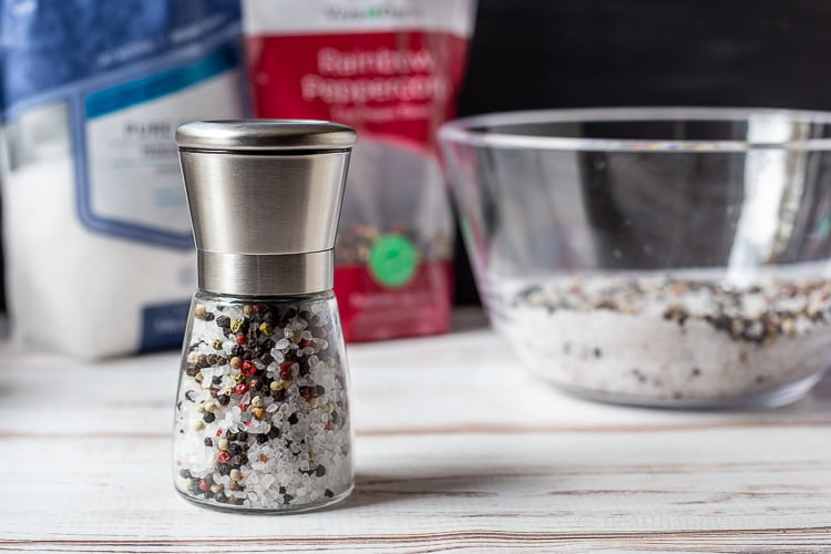 A salt and pepper grinder next to a large clear bowl of the same salt and pepper mix and a bag of salt and a bag of rainbow peppercorns in the background.