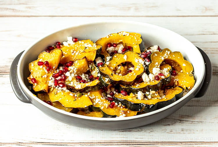 A serving dish with roasted acorn squash slices with pomegranate seeds, pine nuts, pepitos and queso fresco cheese.