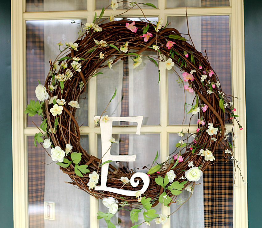Spring wreath with a grapevine base. Pink and light yellow tiny blossom intertwined. A large distressed white chalk painted letter E in the bottom center.