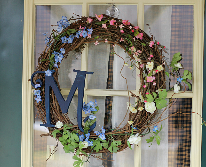 Grapevine wreath with small spring blossoms in pink, blue white and yellow. A large letter M in blue on the side.
