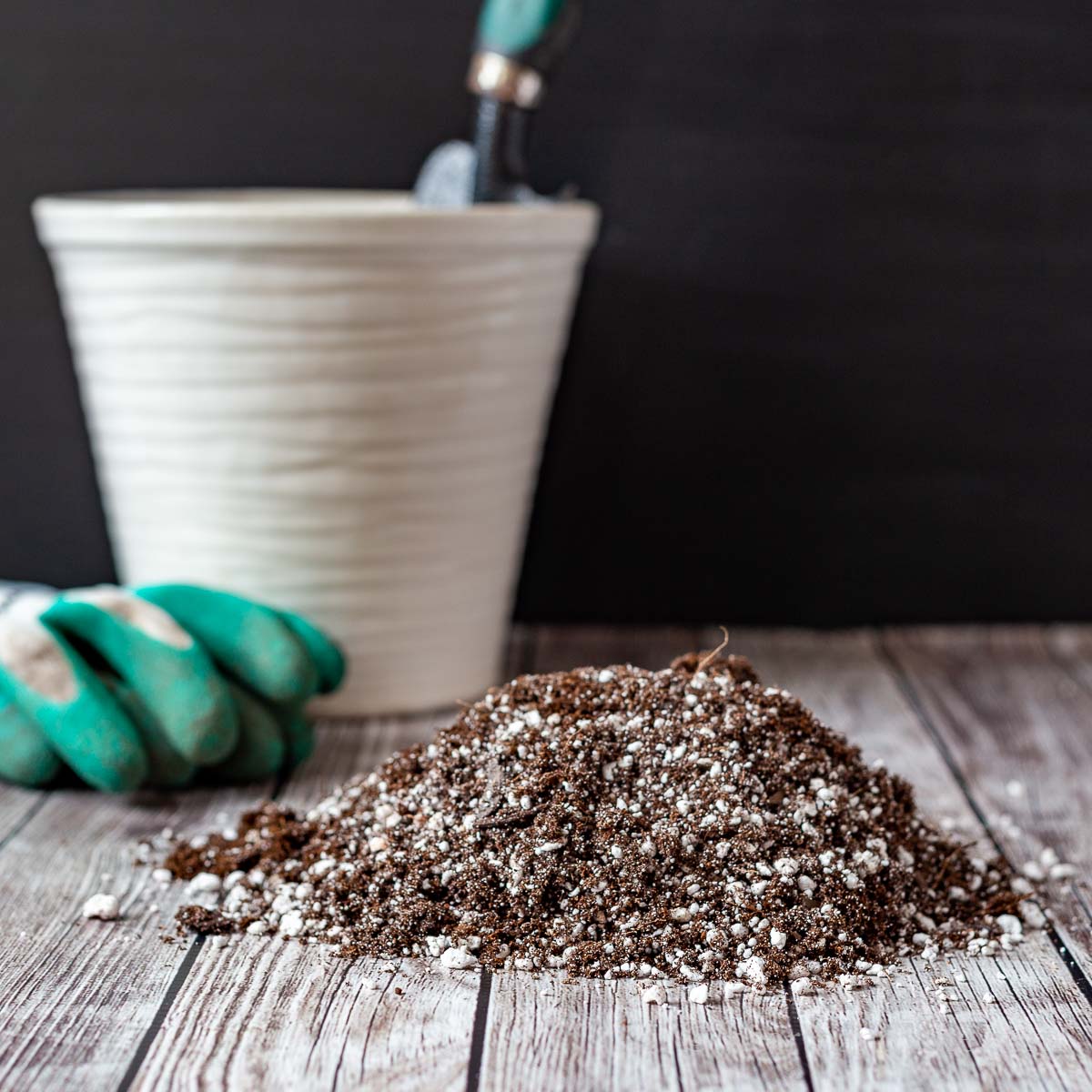 Potting soil on a table next to a white pot and garden gloves.