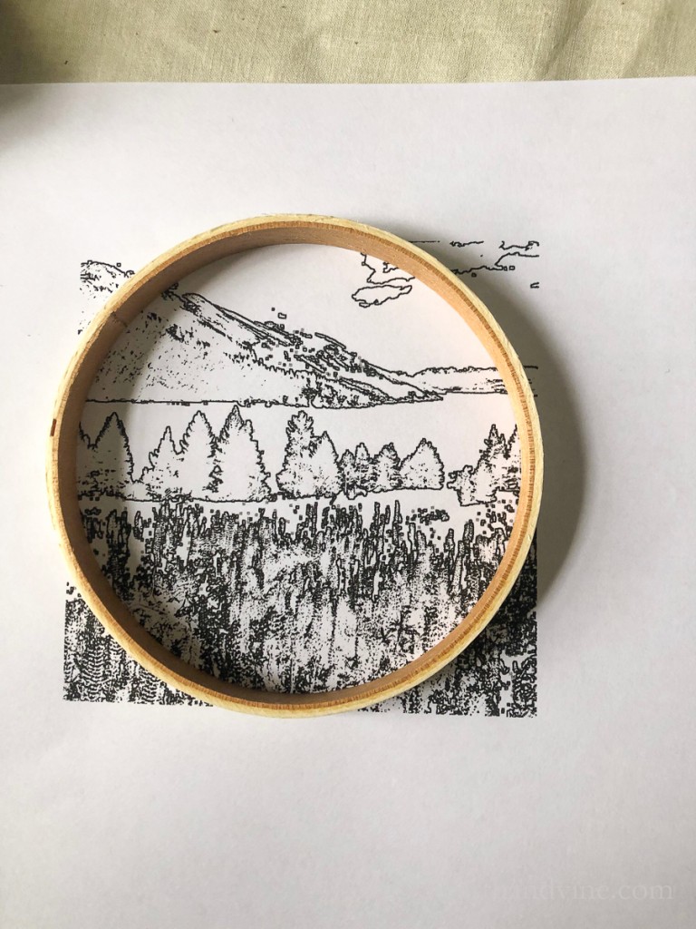 Wooden embroidery hoop covering a black and white landscape drawing.