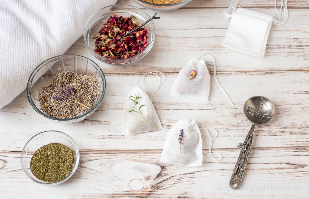 Bowl of peppermint, lavender and rose petals next to a few small white drawstring teabags and a decorative spoon.